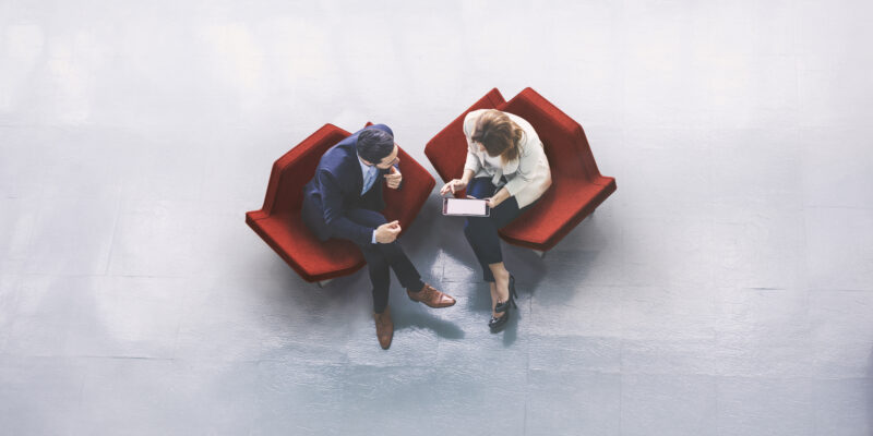 A high-angle view of a businessman and a businesswoman sitting in the office building lobby and using a tablet computer