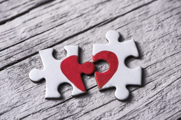 closeup of two separated pieces of a puzzle which together form a heart on a white rustic wooden surface, depicting the idea of rupture or cooperation