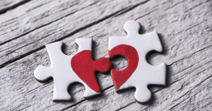closeup of two separated pieces of a puzzle which together form a heart on a white rustic wooden surface, depicting the idea of rupture or cooperation
