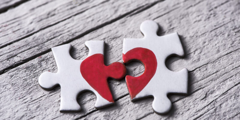 closeup of two separated pieces of a puzzle which together form a heart on a white rustic wooden surface, depicting the idea of rupture or cooperation