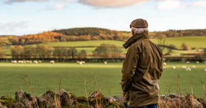 A man with his back to camera wearing a waxed jacket and flat tweed cap looking across a field