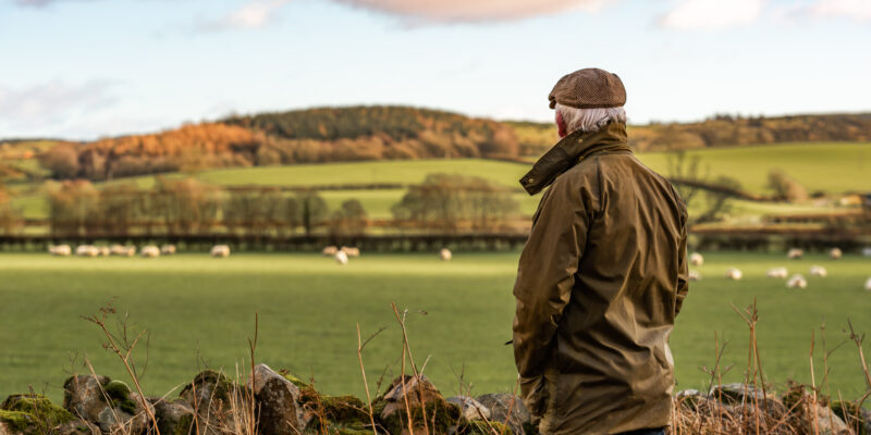 A man with his back to camera wearing a waxed jacket and flat tweed cap looking across a field